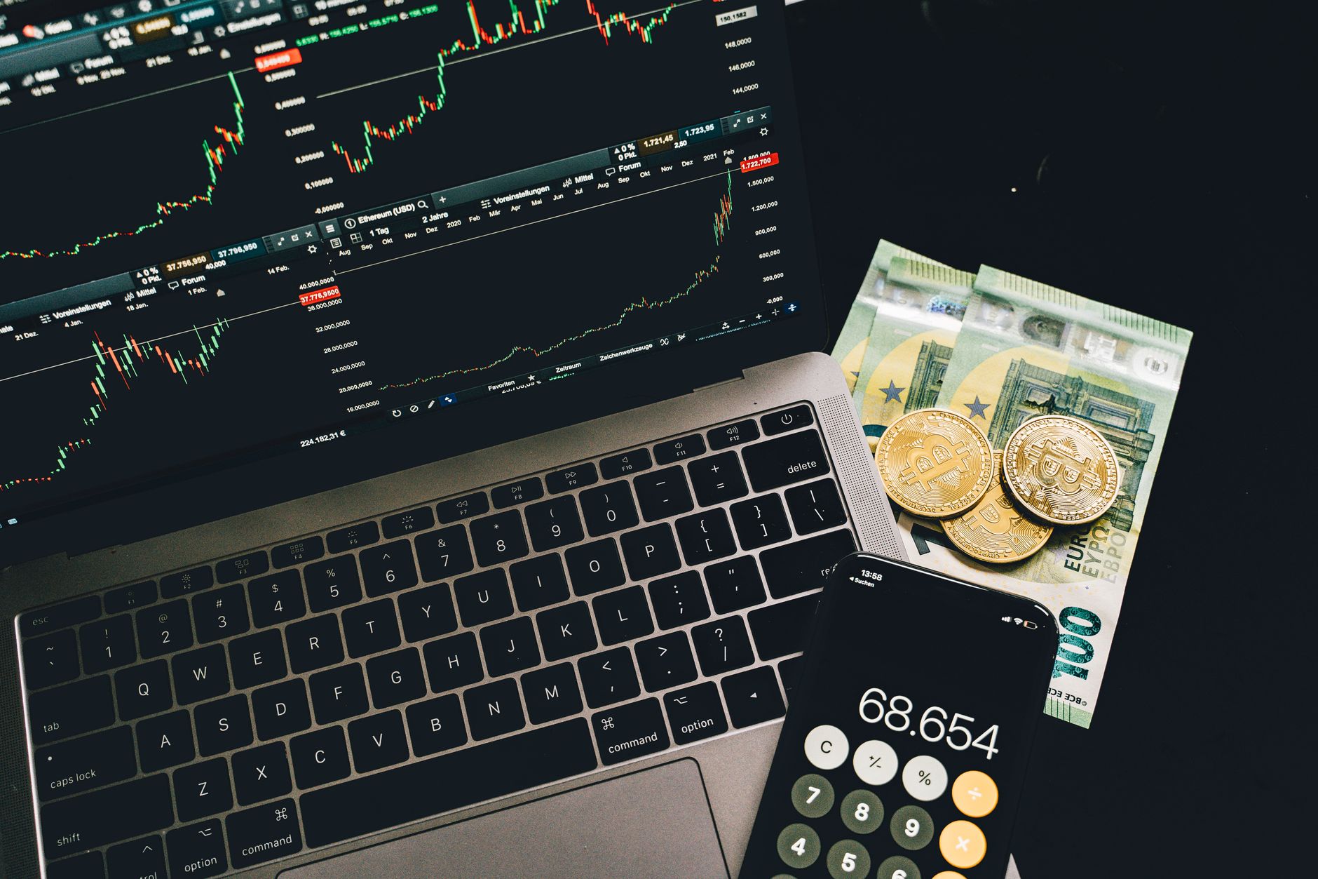 Ordinateur portable affichant des graphiques financiers, billets en euros, pièces dorées et smartphone avec une calculatrice, sur un fond sombre.