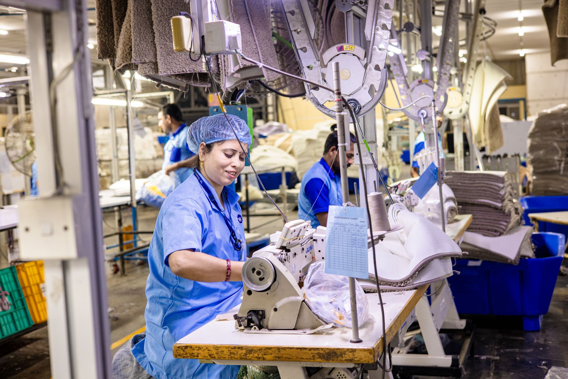 Ouvrière en blouse bleue à une machine à coudre industrielle dans un atelier textile, avec des tapis empilés et d’autres employés au fond.