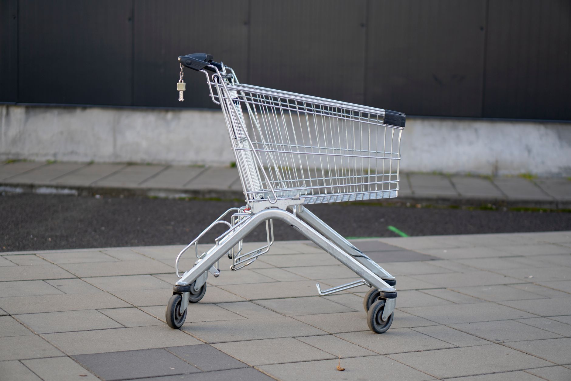 Un caddie de supermarché vide en métal argenté posé sur un sol dallé gris, devant un mur sombre.